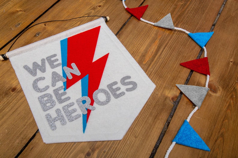 May include: A white felt banner with the text "WE CAN BE HEROES" in silver glitter letters. A red and blue lightning bolt design is behind the text. The banner is hanging from a wooden dowel rod with black string.
