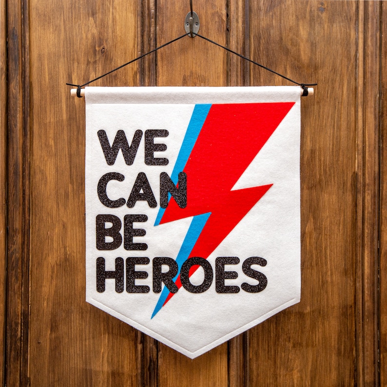 May include: White felt banner with a red and blue lightning bolt design and the text "WE CAN BE HEROES" in black glitter lettering.