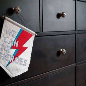 May include: A white felt banner with a red and blue lightning bolt design. The banner reads "We Can Be Heroes" in silver glitter lettering. The banner is hanging on a black dresser with silver knobs.