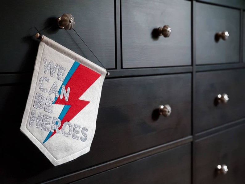 May include: A white felt banner with a red and blue lightning bolt design. The banner reads "We Can Be Heroes" in silver glitter lettering. The banner is hanging on a black dresser with silver knobs.