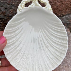 May include: Off-white seashell-shaped dish with a textured, ridged surface. The dish has a scalloped edge with a decorative, light-yellow trim. The dish is held by a person with pink nail polish.