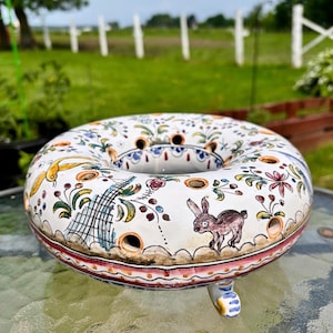 Peut inclure: Anneau décoratif en céramique blanche peint à la main avec des motifs floraux et animaliers. L'anneau présente un lapin, des oiseaux et des fleurs de différentes couleurs. Il comporte plusieurs trous et un pied décoratif.