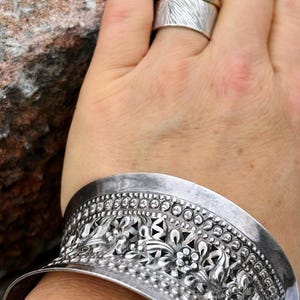 May include: A silver bracelet with intricate floral designs and a textured surface. The bracelet is wide and has a curved shape.