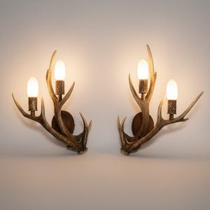 Può includere: Due applique a forma di corno di cervo con lampadine a luce calda. Gli applique sono di colore marrone rustico, montati su piastre posteriori circolari marrone scuro. Il design evoca un'estetica naturale, in stile rustico.