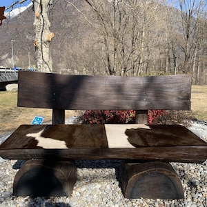 Può includere: Una panca in legno rustica con una seduta in pelle di mucca. La panca è realizzata con tronchi grezzi e ha una finitura naturale e invecchiata. La seduta è realizzata in pelle di mucca nera e bianca con un design a patchwork. La panca è posizionata sullo sfondo di alberi e una catena montuosa.