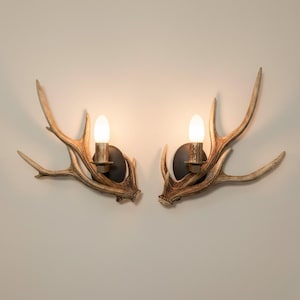May include: A pair of wall-mounted light fixtures featuring natural-colored deer antlers as the primary structure. Each fixture includes a dark circular base, a metallic candle-style socket, and a single glowing white light bulb.