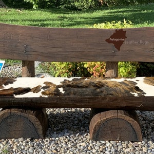 May include: A rustic wooden bench with a cowhide seat. The bench has a wooden back and two large wooden legs. The bench is sitting on a bed of gravel. The bench has a sign on the back that says "Patagonia Leather Rugs".