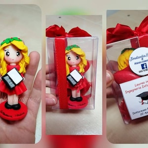 Può includere: Una piccola figurina rossa e bianca di una ragazza con un vestito rosso e una corona verde che tiene un computer portatile. La figurina è in una scatola di plastica trasparente con un nastro rosso. La scatola ha un'etichetta che dice "Laurea in Ingegneria Informatica" e "Dettoressa Nadia Orlandi 22 Aprile 2022".