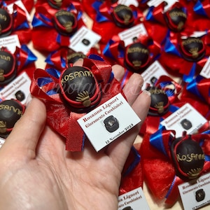 Può includere: Primo piano di un fiocco rosso e blu con una piccola figurina nera e dorata di un cappello militare. La figurina è attaccata a un cartoncino bianco con il testo "Rosanna Liguori Giuramento Carabinieri 13 Novembre 2023".