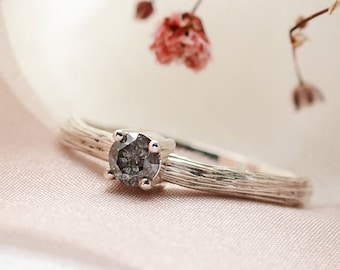 May include: A silver ring with a gray diamond set in a textured band. The ring is displayed on a white and pink fabric background.
