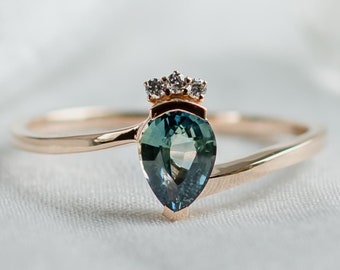 May include: A gold ring with a pear-shaped, teal-colored gemstone. The ring features a small crown design above the gemstone, with three small, clear stones. The ring is set on a white surface.