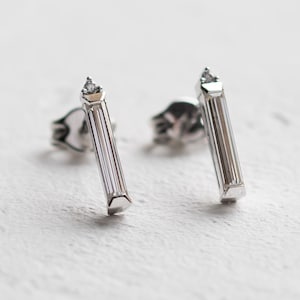 May include: A pair of silver stud earrings. Each earring features a vertical, rectangular design with a small, clear gemstone at the top. The earrings are set against a white, textured background.