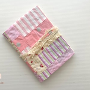 Può includere: Una copertina per quaderno patchwork con una varietà di tessuti rosa e bianchi, tra cui righe, quadretti e motivi floreali. La copertina è rifinita con pizzo color crema, creando un design delicato e femminile.