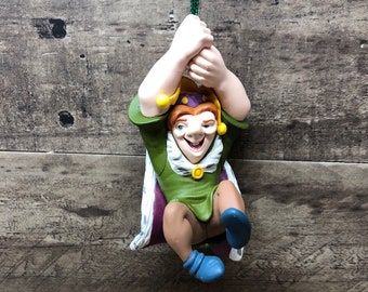 Può includere: Una statuetta di giullare verde e viola con un cappello giallo e un colletto bianco. Il giullare è appeso a una corda verde e ha un viso sorridente.