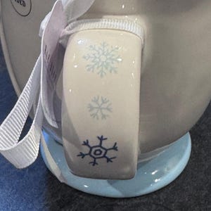 Puede incluir: Una taza de cer&aacute;mica blanca con un dise&ntilde;o de copo de nieve azul en el asa. La taza tiene una base azul claro y una cinta blanca.