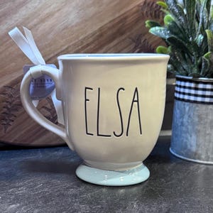 Puede incluir: Una taza de cer&aacute;mica blanca con un borde y una base de color azul claro. La taza tiene el nombre "Elsa" escrito en negro en la parte delantera.