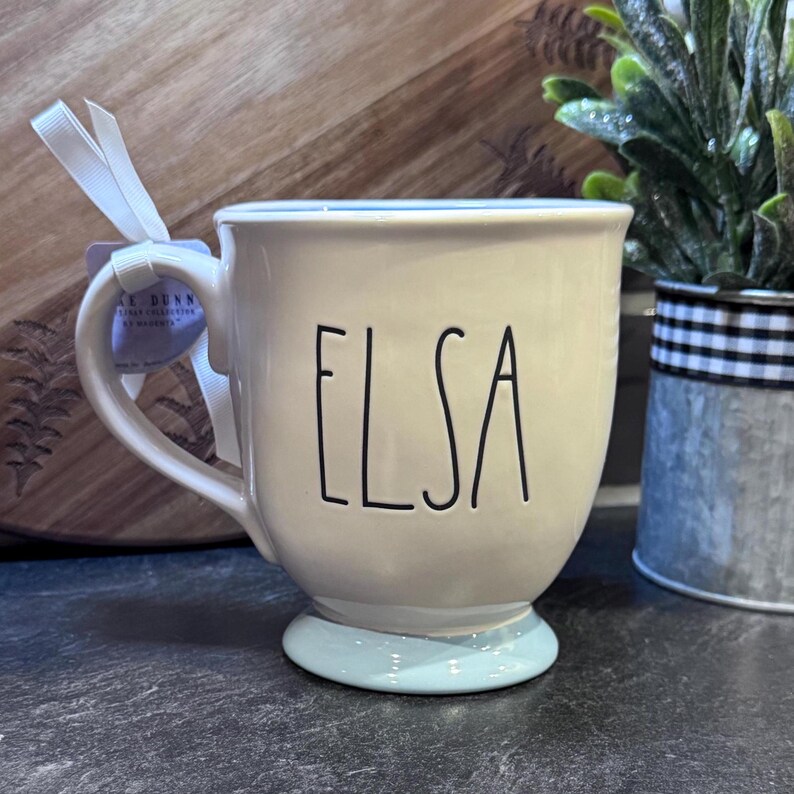 Puede incluir: Una taza de cer&aacute;mica blanca con un borde y una base de color azul claro. La taza tiene el nombre "Elsa" escrito en negro en la parte delantera.