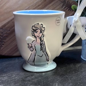 Puede incluir: Taza de cer&aacute;mica blanca con interior azul y borde azul. La taza presenta una ilustraci&oacute;n dibujada a mano de Elsa de Frozen, con el texto "Double Sided" en el lateral.