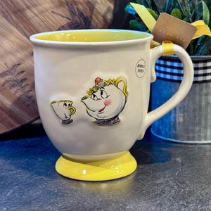May include: A white ceramic mug with a yellow rim and base. The mug features a cartoon illustration of Mrs. Potts and Chip from Disney's Beauty and the Beast. The mug has a double-sided design.