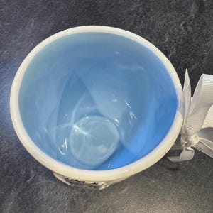 Puede incluir: Una taza de cer&aacute;mica blanca con un interior azul claro. La taza tiene una forma redondeada y un acabado liso y brillante.