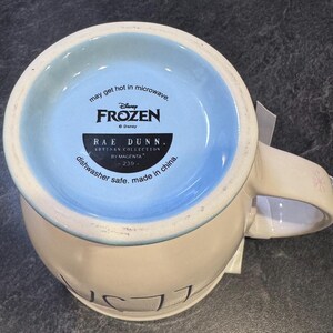 Puede incluir: Una taza de cer&aacute;mica blanca con un borde azul y un dise&ntilde;o de Frozen. La taza tiene el texto "may get hot in microwave, Disney Frozen, Rae Dunn. Artisan Collection by Magenta 239- dishwasher safe. made in china." en la parte inferior.