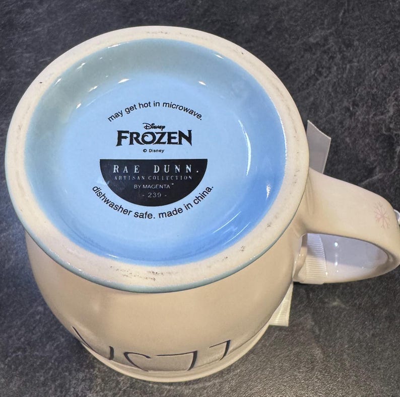 Puede incluir: Una taza de cer&aacute;mica blanca con un borde azul y un dise&ntilde;o de Frozen. La taza tiene el texto "may get hot in microwave, Disney Frozen, Rae Dunn. Artisan Collection by Magenta 239- dishwasher safe. made in china." en la parte inferior.
