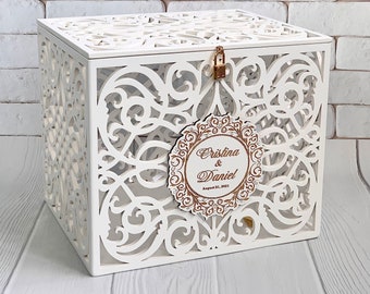 May include: White wooden box with a decorative, intricate, cut-out design. The box has a lock and keyhole. A gold circle with the text "Cristina & Daniel August 21, 2021" is attached to the box.