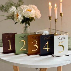 Op de afbeelding: Vijf tafelnummers in verschillende kleuren: bruin, groen, tan, zwart en wit. Elk bord heeft een gouden nummer en het woord "tafel" in elegant schrift. De borden staan op een witte tafel met kaarsen en bloemen op de achtergrond.