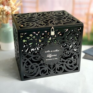 May include: Black wooden wedding card box with a decorative, intricate, cut-out design. The box has a lock and key and a personalized label that reads "Mr. & Mrs. Lyons November 11, 2024".