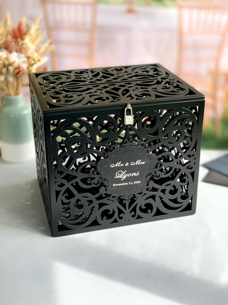 May include: Black wooden wedding card box with a decorative, intricate, cut-out design. The box has a lock and key and a personalized label that reads "Mr. & Mrs. Lyons November 11, 2024".