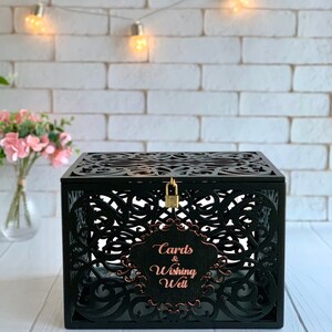 May include: Black wooden box with intricate cutouts and a lock. The box has a sign that says "Cards & Wishing Well" in rose gold lettering.