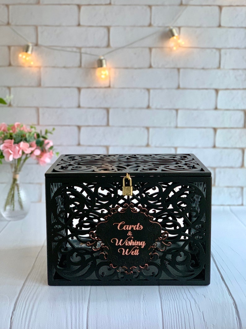 May include: Black wooden box with intricate cutouts and a lock. The box has a sign that says "Cards & Wishing Well" in rose gold lettering.