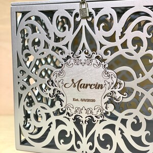 May include: Silver metal box with a decorative, intricate cut-out design. The box has a lock and a wooden plaque with the text "Marcin Est. 8/8/2020".