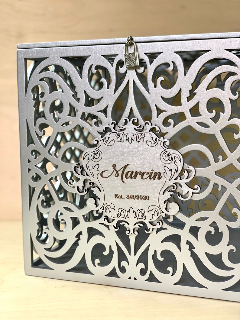 May include: Silver metal box with a decorative, intricate cut-out design. The box has a lock and a wooden plaque with the text "Marcin Est. 8/8/2020".