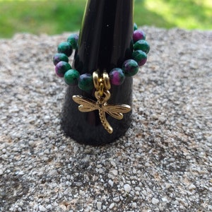 Peut inclure: Une bague en pierre précieuse verte et rose avec un pendentif en forme de libellule dorée. La bague est présentée sur un porte-bague noir.