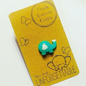 May include: A teal blue elephant figurine with a white heart and a white top. The figurine is on a brown card with the text "Fresh Banana Crafts" and "You Are Unforgettable".