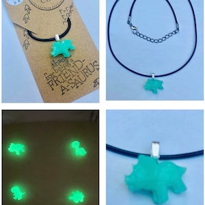 Könnte beinhalten: Eine grüne, im Dunkeln leuchtende Dinosaurier-Anhängerkette auf einem schwarzen Band. Der Anhänger ist in Form eines Triceratops geformt. Die Kette ist auf einer Karte verpackt, auf der "Fresh Banana Crafts" und "Danke, dass du ein Freund-A-saurus bist" steht.