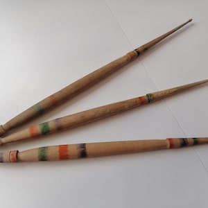 Può includere: Tre ferri da maglia in legno con finitura in legno naturale. Ogni ago ha un'estremità appuntita ed è decorato con fasce colorate, tra cui verde, arancione e viola. Gli aghi sono di lunghezze variabili.