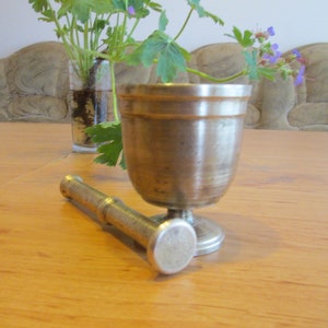 May include: A silver mortar and pestle set on a wooden table. The mortar is a cup-shaped bowl with a rounded base. The pestle is a long, cylindrical rod with a rounded end.