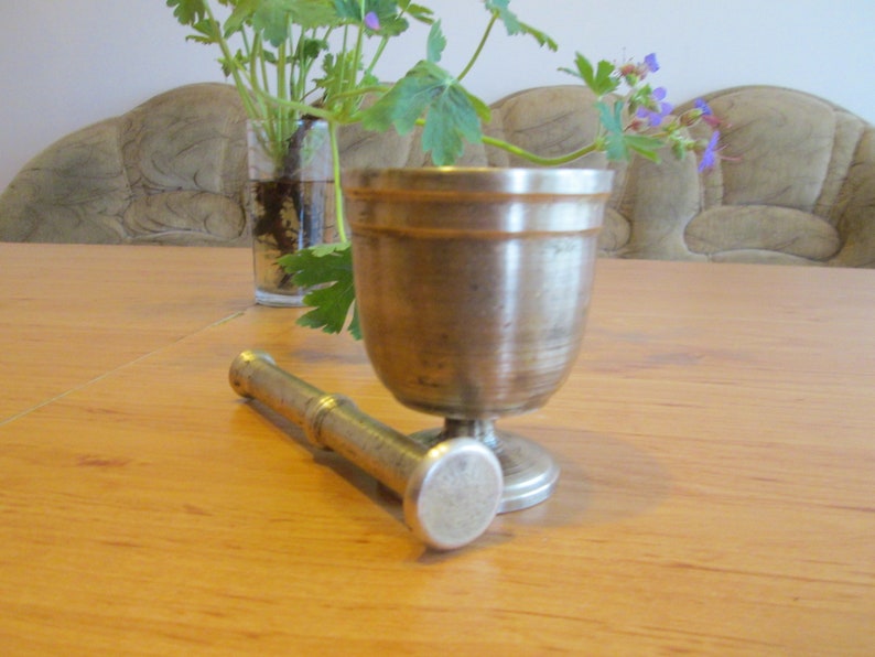 May include: A silver mortar and pestle set on a wooden table. The mortar is a cup-shaped bowl with a rounded base. The pestle is a long, cylindrical rod with a rounded end.