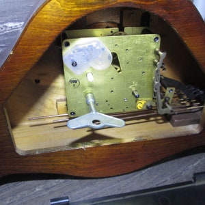 May include: Close-up of the inner workings of a wooden clock. The clock face is missing, but the brass gears and winding key are visible. The clock is made of wood and has a dark brown finish.