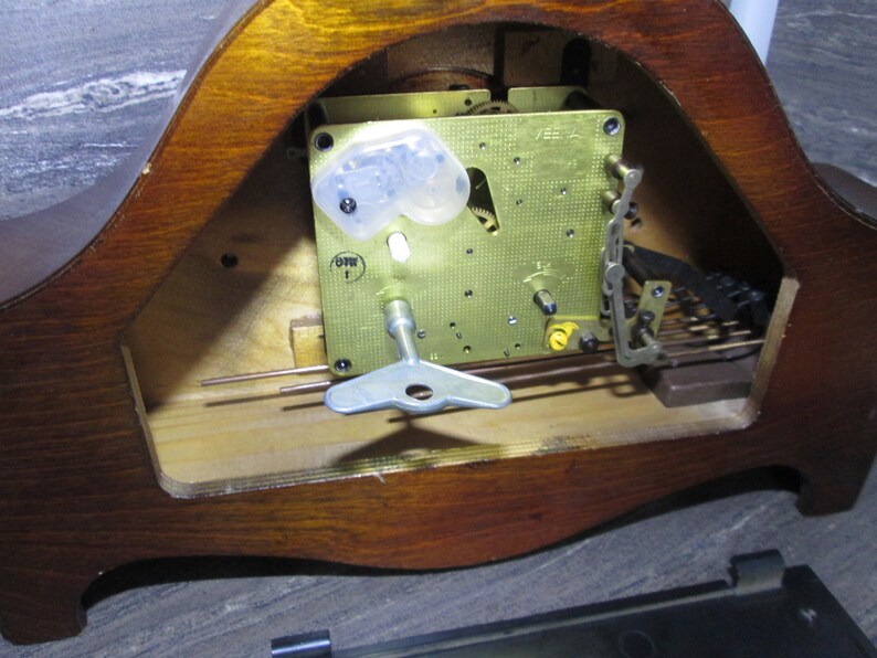 May include: Close-up of the inner workings of a wooden clock. The clock face is missing, but the brass gears and winding key are visible. The clock is made of wood and has a dark brown finish.