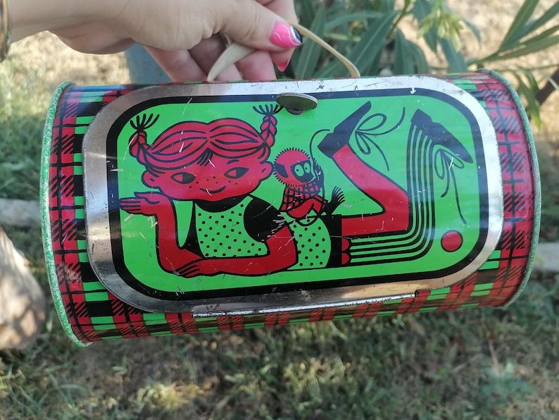 May include: Vintage metal lunchbox with a whimsical illustration of a girl and a monkey. The lunchbox has a red, green, and black plaid pattern on the sides. The main image features a girl with red hair and a monkey on a green background.