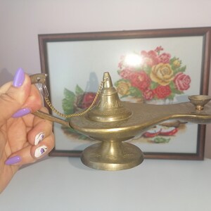 May include: A gold-coloured genie lamp with intricate details and a chain attached to the lid. The lamp is sitting on a white surface.