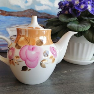 Könnte beinhalten: Eine weiße Porzellan-Teekanne mit goldener Verzierung und einem floralen Muster aus rosa Rosen. Die Teekanne hat einen Deckel und einen Henkel.