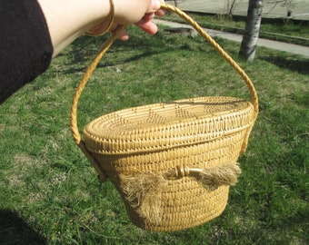 May include: A woven basket with a braided handle and a lid. The basket is oval-shaped and made of light brown material. A decorative tassel hangs from the side. The basket is being held up by a person.