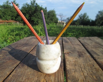May include: A white marble pencil holder with three pencils inside. The pencils are orange, yellow, and black. The holder is sitting on a wooden table.