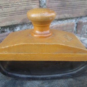 May include: A wooden inkwell with a black metal base and a wooden lid with a knob. The inkwell has a rectangular shape and is made of light brown wood.