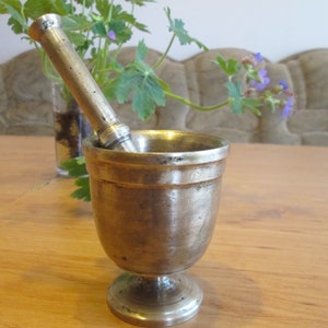 May include: A silver mortar and pestle set on a wooden table. The mortar is a cup-shaped bowl with a wide base and a narrow top. The pestle is a long, cylindrical rod with a rounded end.