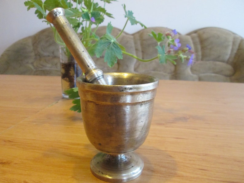 May include: A silver mortar and pestle set on a wooden table. The mortar is a cup-shaped bowl with a wide base and a narrow top. The pestle is a long, cylindrical rod with a rounded end.